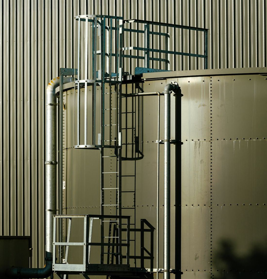 A caged fixed ladder on an industrial storage tank, leading to a guardrailed platform at the top.